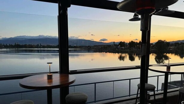 Une terrasse offrant une vue sur un lac paisible et les montagnes au coucher du soleil. On y trouve des tables rondes et des tabourets de bar.