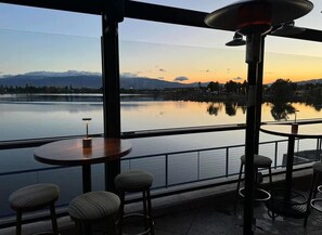 A terrace with a view of a calm lake and mountains at sunset. There are round tables and bar stools. - Amazing Water View w Jacuzzi Sky Lounge (Sparks)