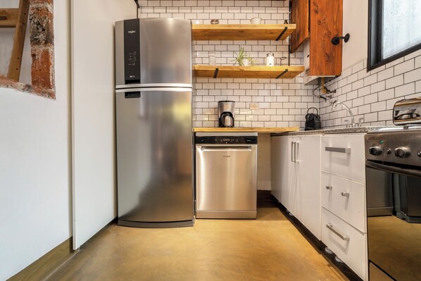 A compact kitchen with a stainless steel refrigerator, dishwasher, and oven. There are wooden shelves above the countertop and a window with a view.