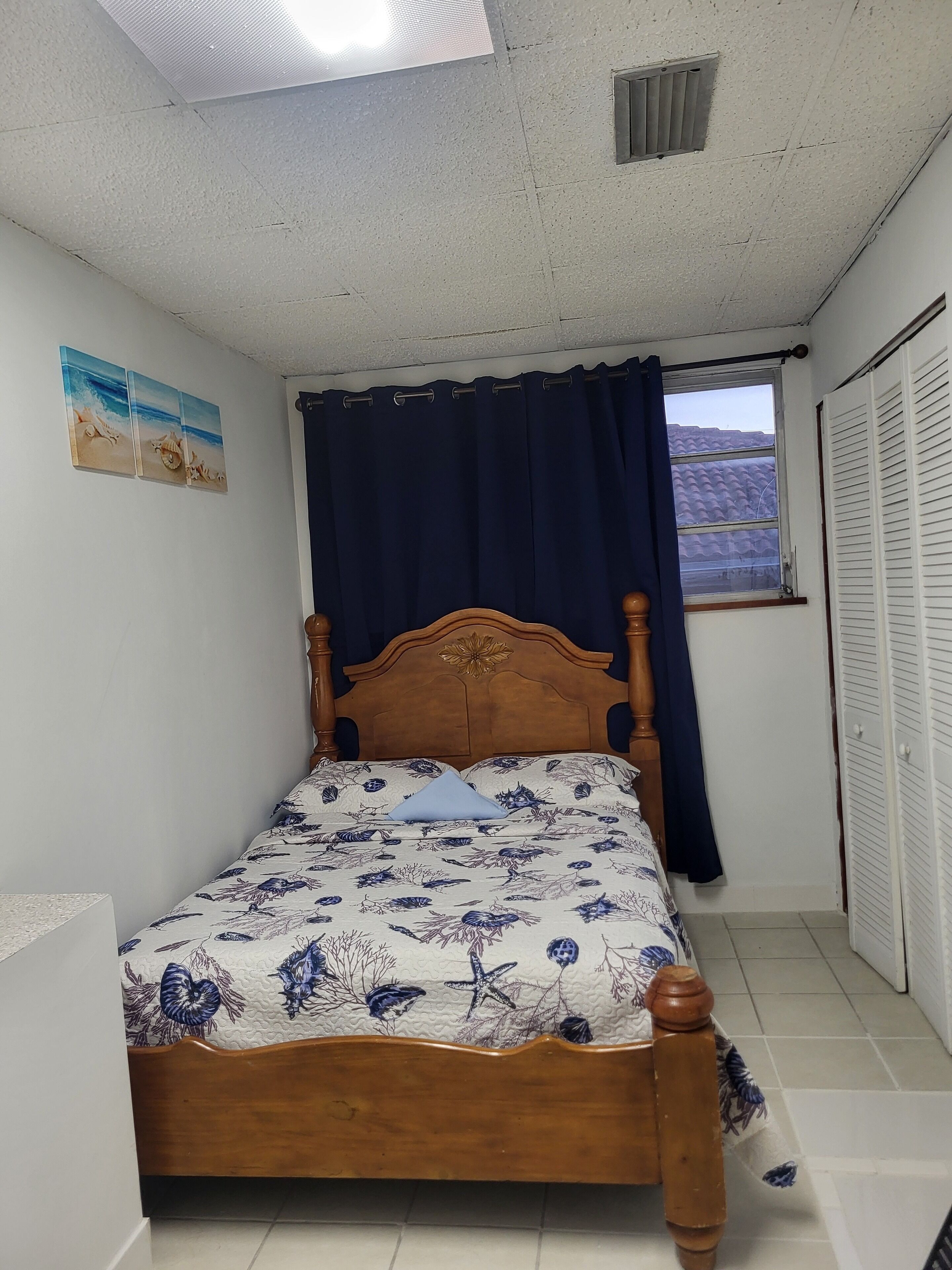 A single bed with a floral bedspread, a wooden headboard, a window with a view of a tiled roof, and a closed white wardrobe.
