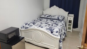 A bedroom with a white bed, a small bedside table, a microwave, and a wall-mounted air conditioner.