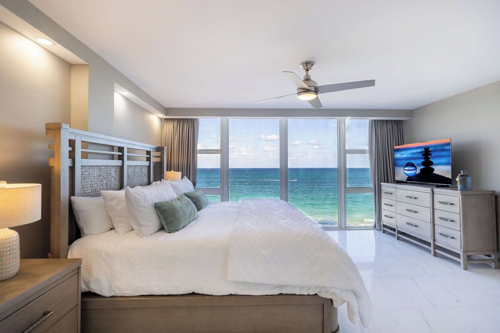 Un dormitorio con una cama grande, ventilador de techo, televisor y vista al océano.