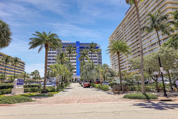Entrada de hotel con un letrero azul, palmeras y un estacionamiento.