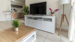 A modern living room with a flat-screen TV on a white entertainment unit, a wooden coffee table with a plant, a standing lamp, and a dining area in the background. - Proche Thalazur et plages, Parking privé, Piscine (Antibes)