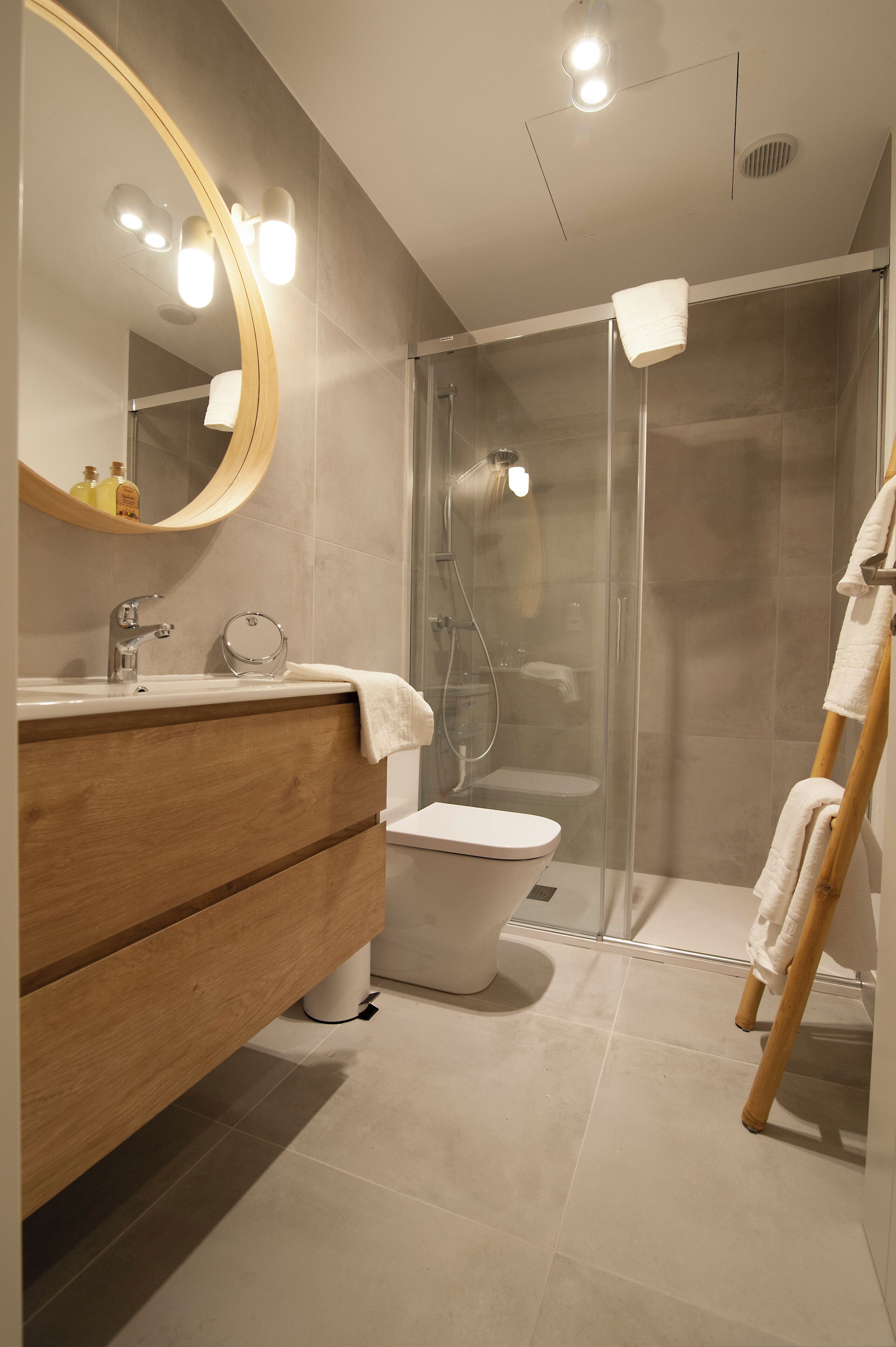 A modern bathroom with a wooden vanity, a round mirror, a glass shower enclosure, and a white toilet.