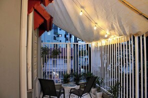 Un área de estar al aire libre con una mesa y sillas, un toldo blanco y vistas de edificios al fondo.