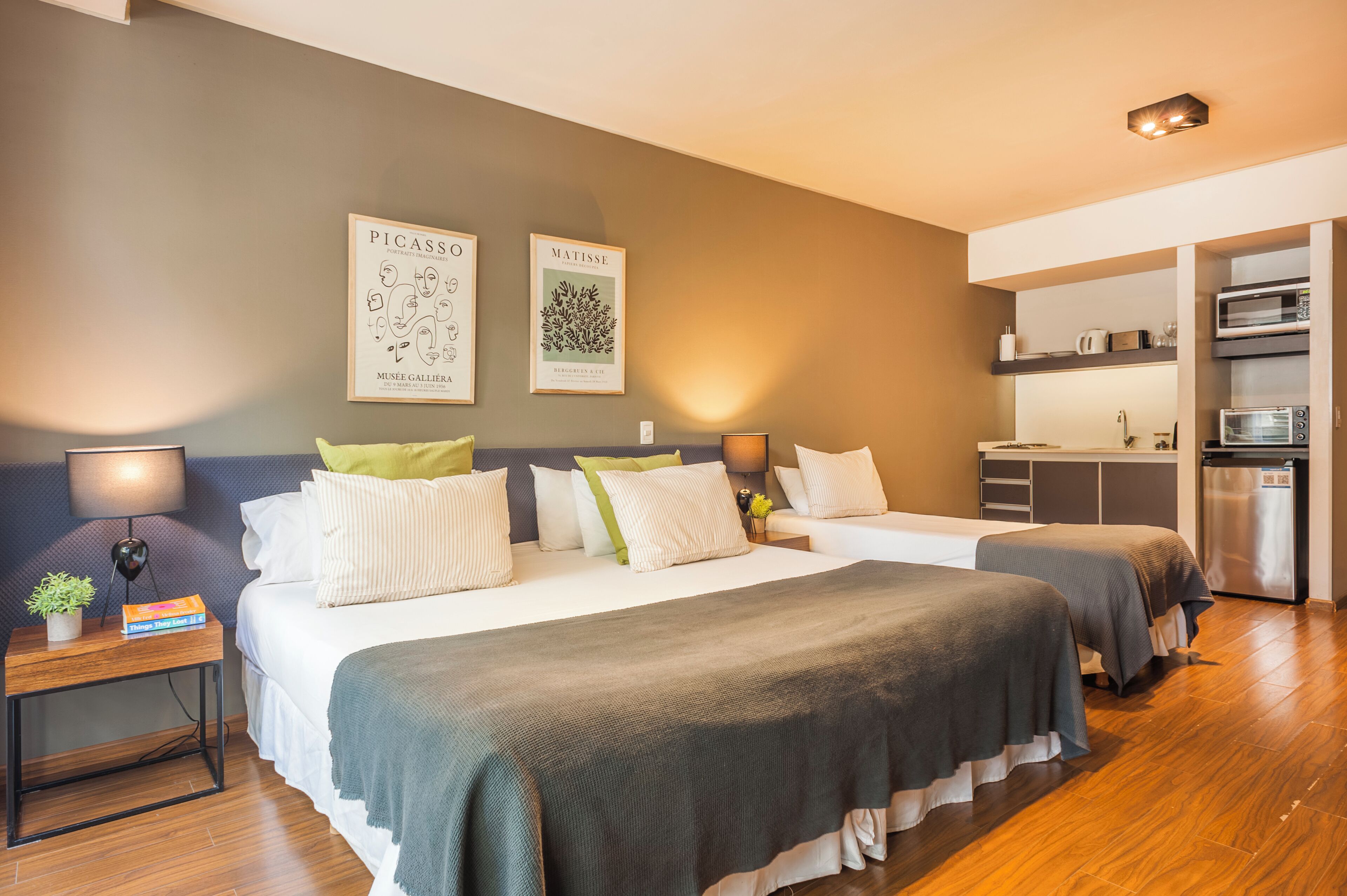 A modern hotel room with two beds, a small kitchenette, and artwork on the wall.