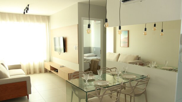 Una sala de estar moderna con una mesa de comedor de cristal, sillas blancas, un sofá y un televisor montado en la pared.