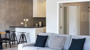 Una sala de estar moderna con un sofá gris, cojines negros estampados y una zona de cocina con un taburete alto y un jarrón con flores.