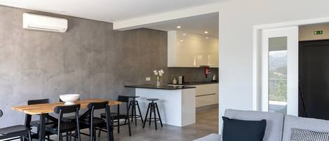 Una sala moderna con un sofá gris, una mesa de comedor de madera con sillas negras, y una cocina con encimera blanca y taburetes de bar.