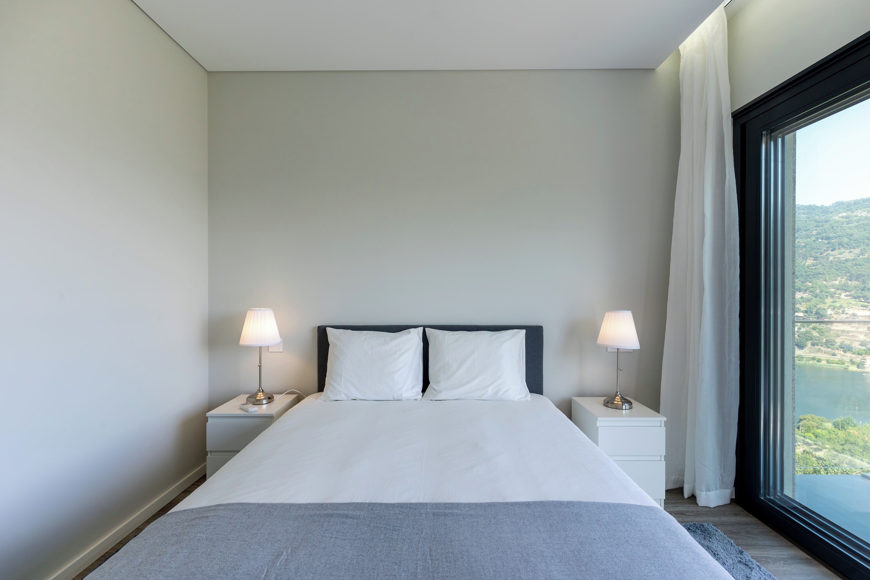 A modern hotel room with a large bed, two bedside tables with lamps, and a view of a lake through a large window.