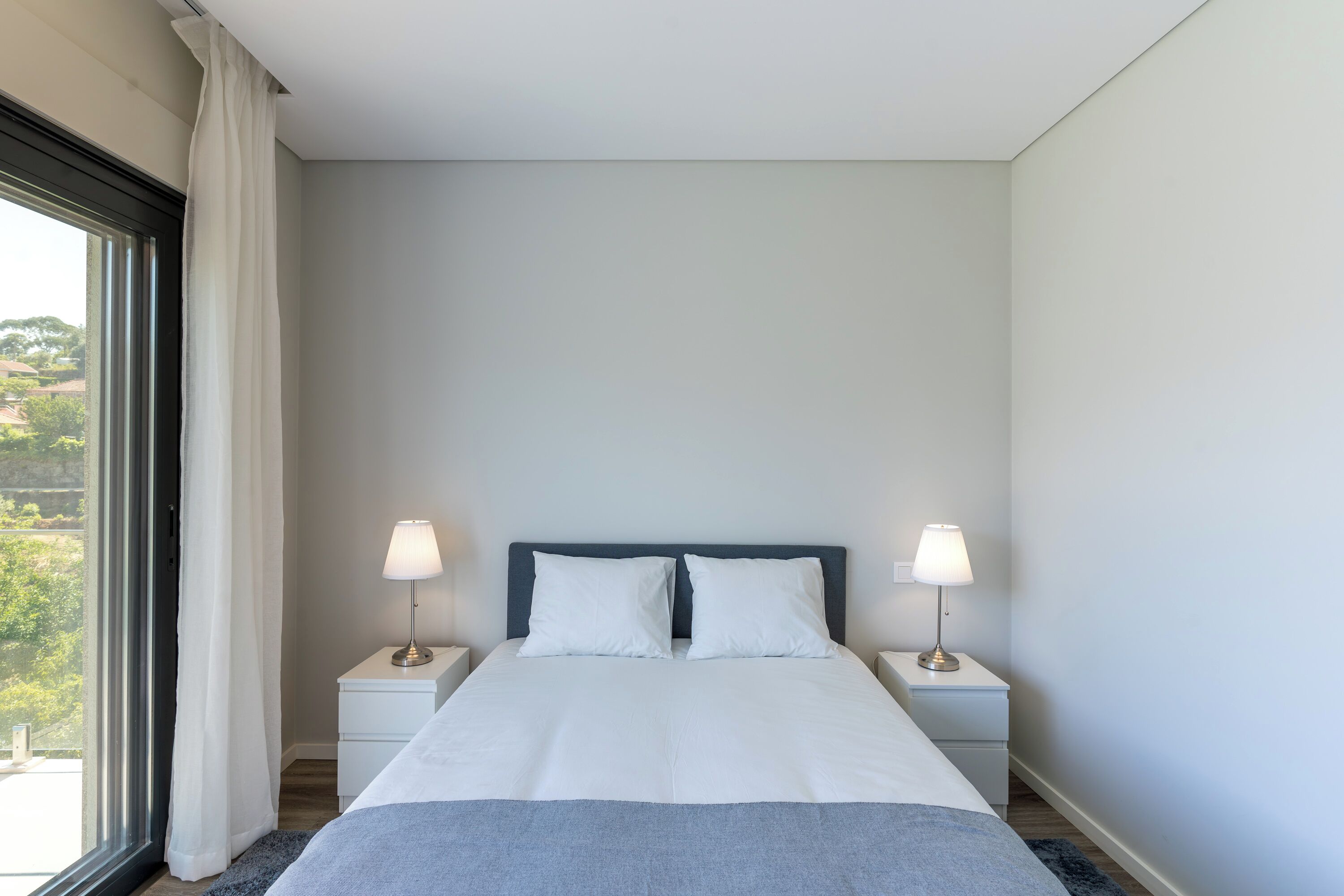 Una cama bien hecha con sábanas blancas, dos lámparas de noche y vistas a zonas verdes a través de una ventana con cortinas blancas.