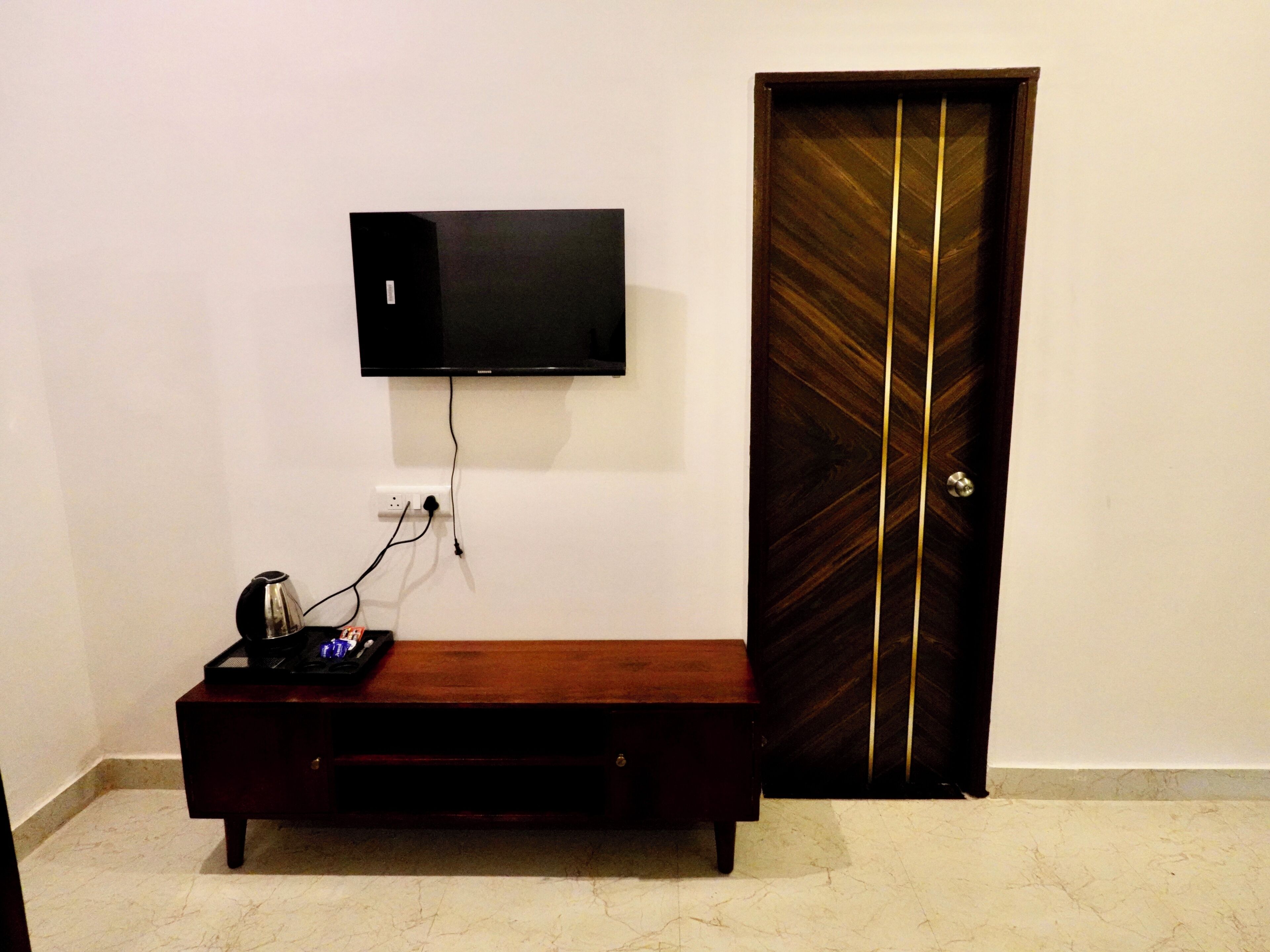 A room with a flat-screen TV mounted on the wall, a wooden bench with a tray and a canister, and a dark wooden door with vertical gold stripes.