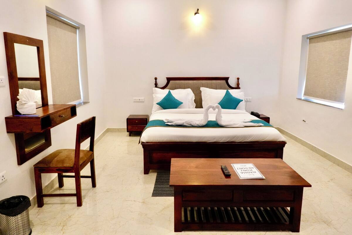 Une chambre d’hôtel comprenant un lit, une table de chevet en bois, un bureau, une chaise, une poubelle et un luminaire fixé au mur.