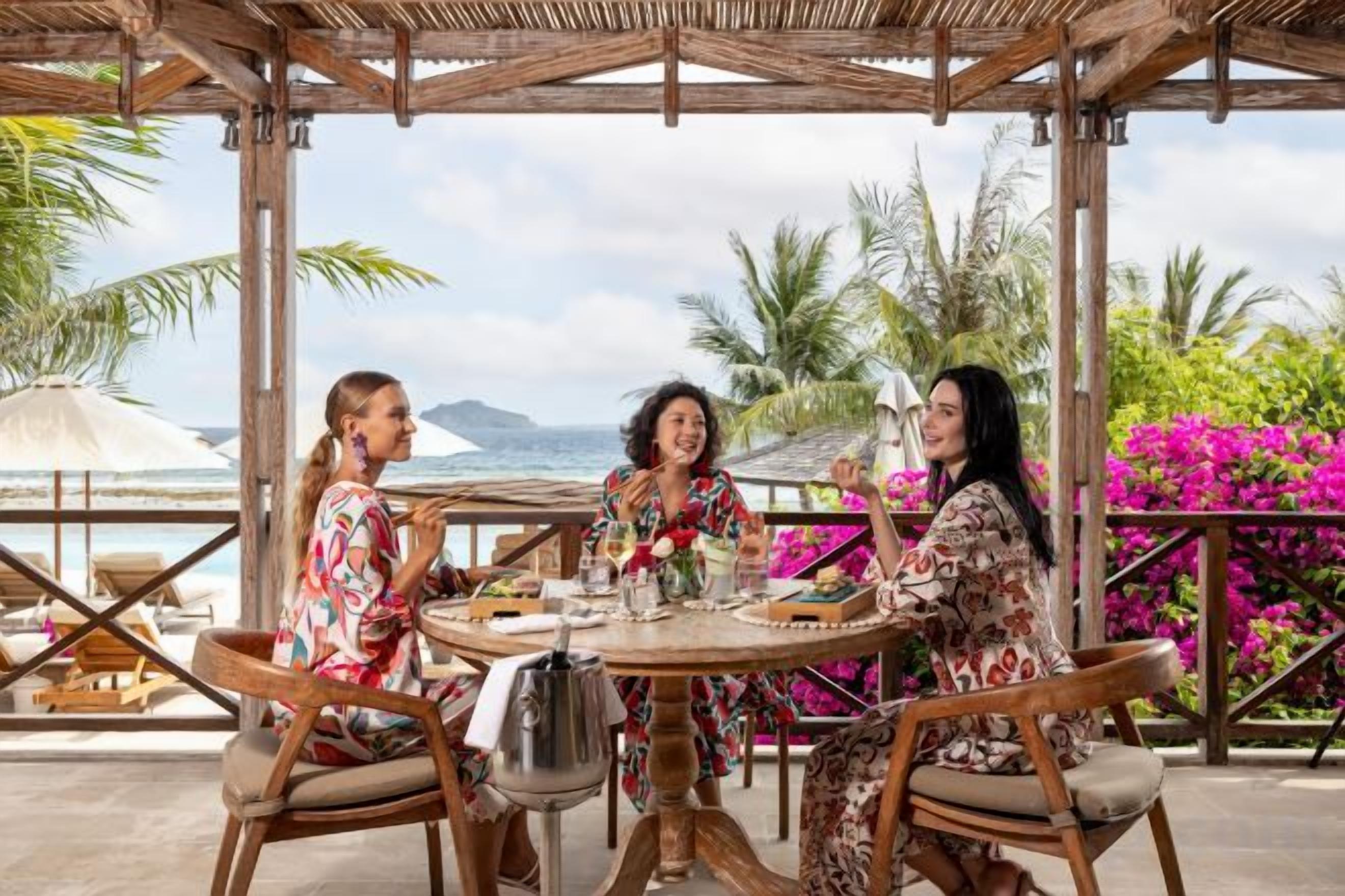 Un groupe de personnes est assis à une table en plein air, devant un paysage offrant une vue sur l’océan et les palmiers.