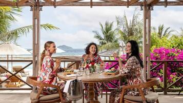 Un groupe de personnes est assis à une table en plein air, devant un paysage offrant une vue sur l’océan et les palmiers.