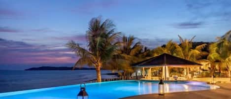 A pool area with a view of the sea, palm trees, and a building with a roof.