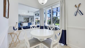 A dining area with a round white table and chairs, a chandelier, and a view of the outdoors through large windows.