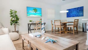 A modern living room with a wooden coffee table, a TV displaying a beach scene, a dining area with wooden chairs, and a large blue abstract painting. - New! Fun in the Sun - Beautifully Renovated! Heated Pool - Pool Table! (Naples)