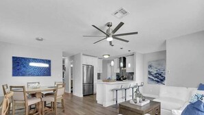 A modern living room with a white sofa, a dining area with wooden chairs, a kitchen with white cabinets, and a ceiling fan. - New! Fun in the Sun - Beautifully Renovated! Heated Pool - Pool Table! (Naples)
