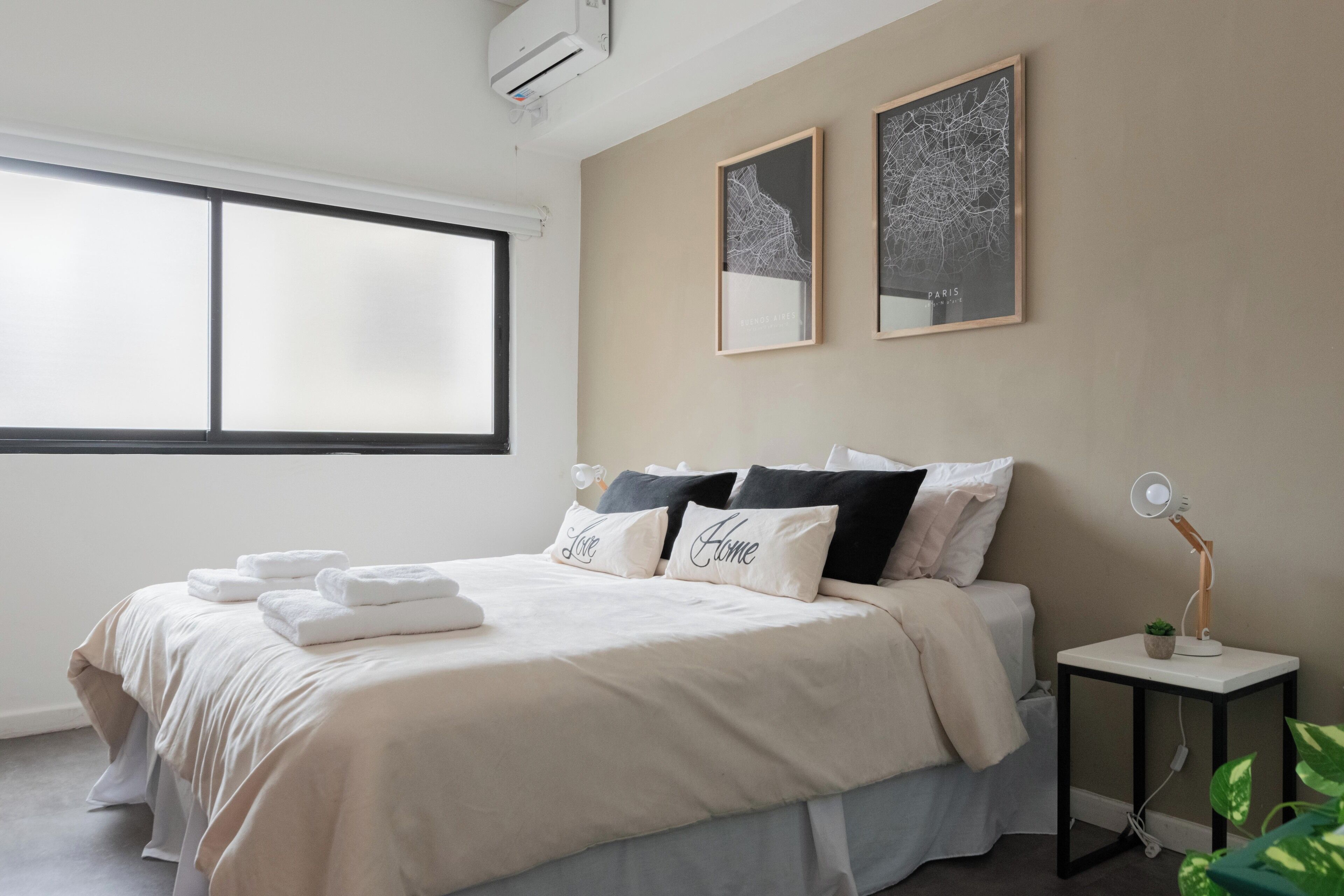 A neatly made bed with white and black pillows, a bedside table with a lamp, and a large window providing natural light.