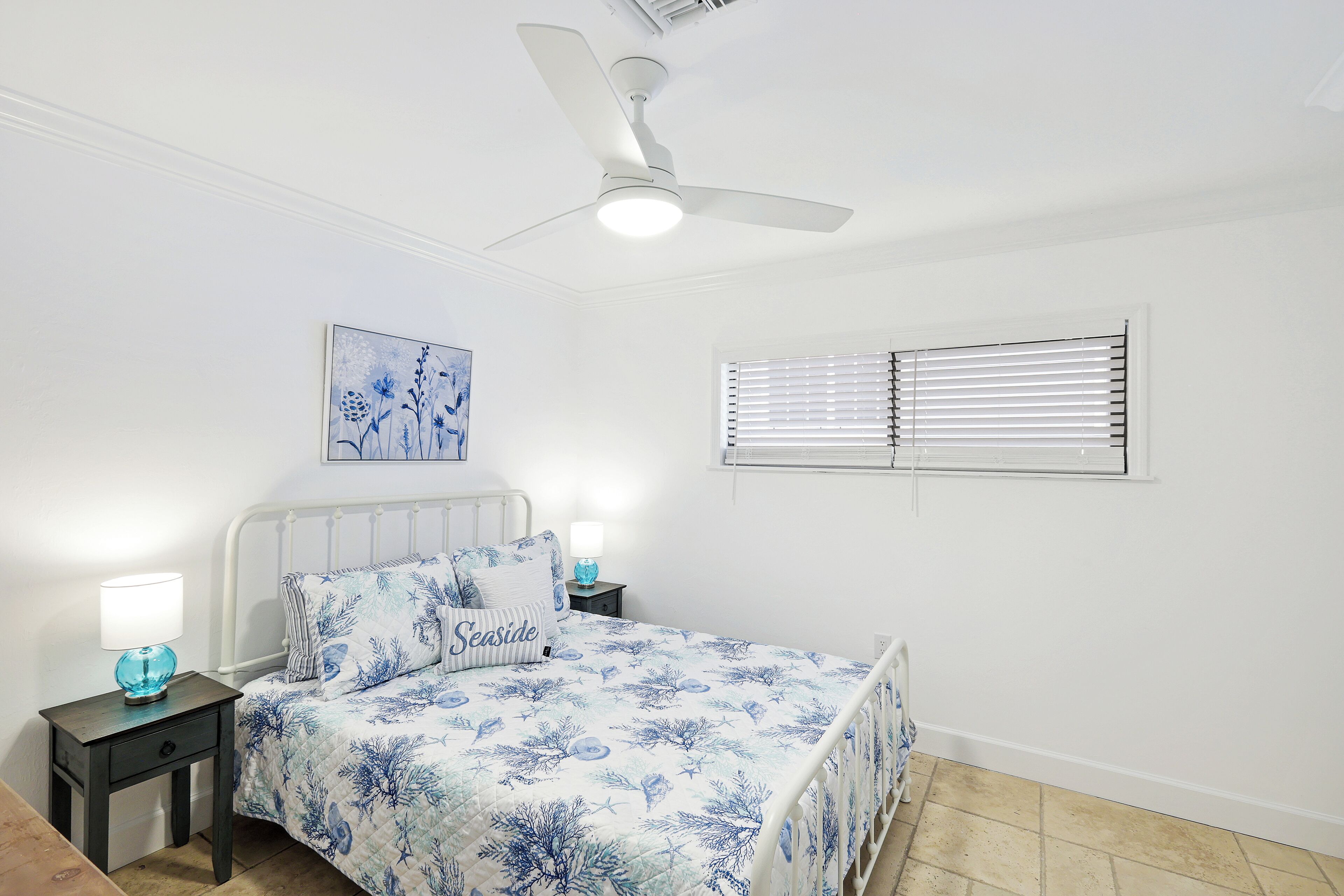 Un dormitorio con cama, mesitas de noche, ventilador de techo, ventana con persianas y un cuadro colgado en la pared.