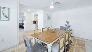 A dining area with a wooden table and six chairs, a ceiling fan, and a kitchen in the background. - Sea La Vie - Pickleball close by, Heated Pool, Walk to Beach! (Naples)