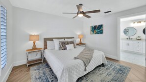 A bedroom with a bed, two bedside tables with lamps, a ceiling fan, a large window with blinds, a bathroom with a mirror and sink, and a blue and white patterned rug.