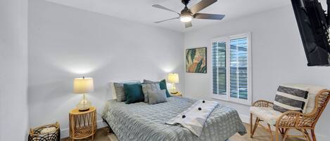 Un dormitorio con cama, dos mesitas de noche con lámparas, un ventilador de techo, una ventana con persianas, una silla y un televisor de pared.