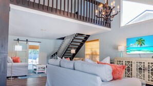 A spacious living room with a large light blue sofa, a wooden staircase, and a mounted TV displaying a tropical beach scene.