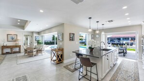 A spacious kitchen with a large island, dining area with chairs, and a view of a swimming pool and boats. - Sunny Side! – Southern Exposure - Heated Saltwater Pool! Walk to Beach! (Naples)