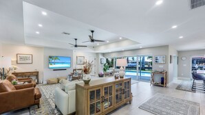 A spacious living room with a large TV, comfortable seating, a wooden cabinet, and a view of the outdoors. - Sunny Side! – Southern Exposure - Heated Saltwater Pool! Walk to Beach! (Naples)