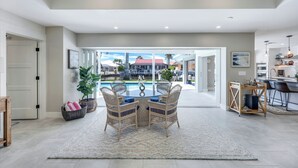 A spacious living area with a dining table, wicker chairs, a large rug, and a view of a pool and outdoor area. - Sunny Side! – Southern Exposure - Heated Saltwater Pool! Walk to Beach! (Naples)