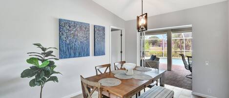 A dining area with a wooden table, chairs, and a bench. There are abstract paintings on the wall, a potted plant, and a hanging light fixture.