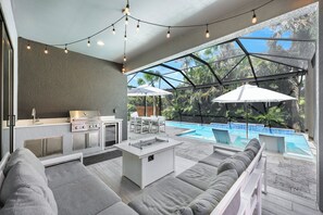 A modern outdoor kitchen with a grill, dining area, and a poolside seating area with umbrellas. - New! Vitamin Sea - 4 Bedroom w/ Heated Salt Water Pool! Walk to Beach! (Naples)