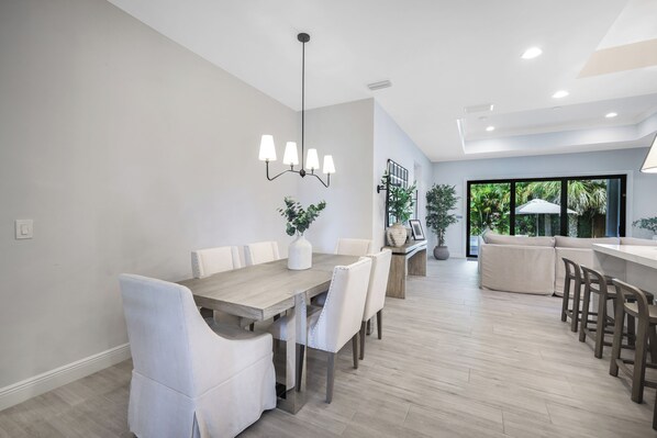 A modern dining area with a wooden table and white upholstered chairs, a hanging light fixture, and a view of a balcony with plants. - New! Vitamin Sea - 4 Bedroom w/ Heated Salt Water Pool! Walk to Beach! (Naples)