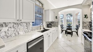 A modern kitchen with white cabinetry, a marble backsplash, and a dining area with a table and chairs. - New! Sunkissed - 1 Mile to Beach! Heated Pool! (Naples)