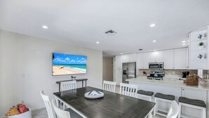 A modern kitchen with white cabinets, a dining area with a dark wooden table and white chairs, and a large flat-screen TV on the wall. - New Listing! Starfish Cove - Heated Pool, Walk to the Beach! (Naples)