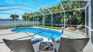 A covered outdoor swimming pool with a glass enclosure, surrounded by wicker chairs and a table with drinks. - New Listing! Starfish Cove - Heated Pool, Walk to the Beach! (Naples)
