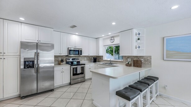 A modern kitchen with white cabinetry, stainless steel appliances, a large island with bar stools, and a window with a view of greenery. - New Listing! Starfish Cove - Heated Pool, Walk to the Beach! (Naples)