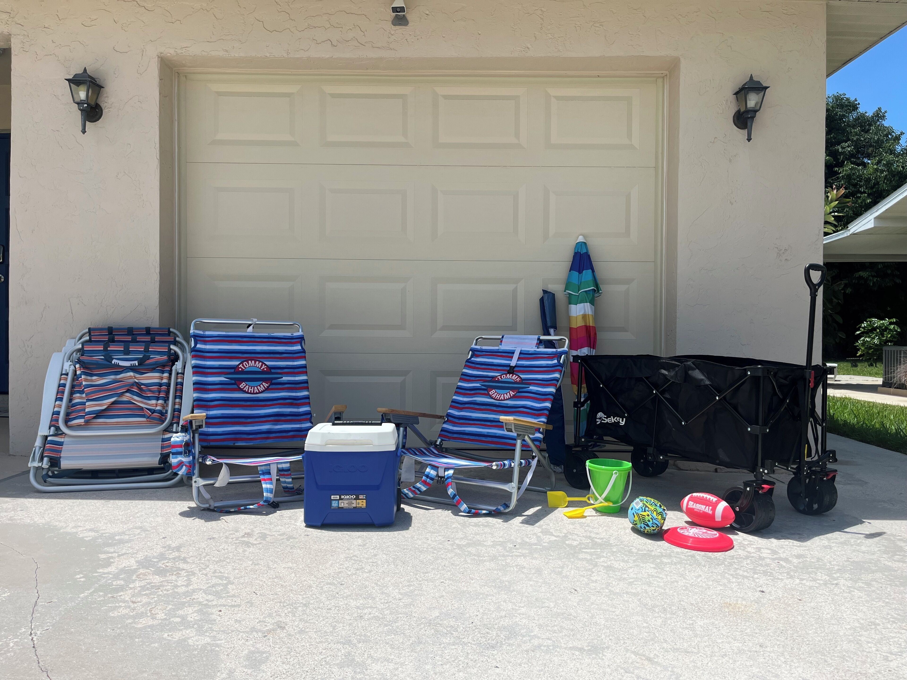 Two beach chairs, a cooler, a beach ball, a bucket, and a stroller in front of a garage door.