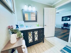 Une salle de bain avec un meuble-lavabo en bois foncé, un miroir avec éclairage intégré, une porte blanche et un couloir menant à une salle à manger.