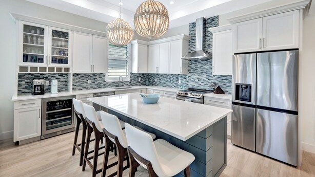 Een moderne keuken met witte kasten, een groot eiland, roestvrijstalen apparatuur en decoratieve verlichting.