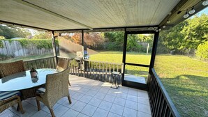Une terrasse couverte avec une table en verre et des chaises en osier, donnant sur une cour arrière équipée d’un barbecue et d’une pelouse.