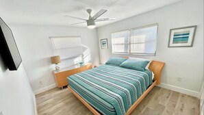 Quarto com cama, mesa de cabeceira, luminária, ventilador de teto e duas janelas com persianas.