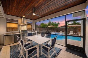 Una cocina exterior con una parrilla, una mesa de comedor y sillas, una piscina con sillas de descanso y vistas al atardecer.