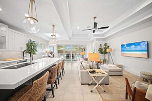 Eine moderne Küche mit einer zentralen Kochinsel, Essbereich mit Korbstühlen, einer Couch und einem Fernseher, der eine Strandansicht zeigt.