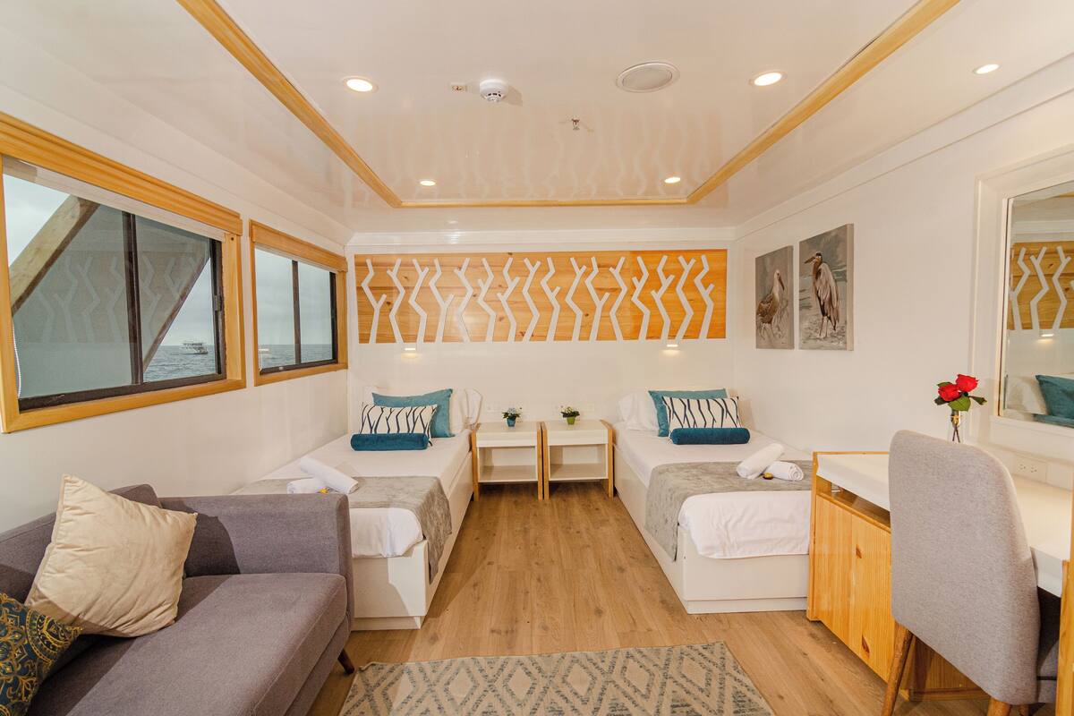 Quarto compacto com duas camas, um sofá, uma escrivaninha e uma cadeira. Há janelas com vista para o mar, uma parede estampada e um espelho.