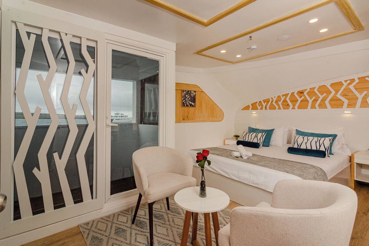 Quarto de hotel moderno com cama, duas cadeiras, uma mesa pequena com um vaso e vista através de uma porta de vidro.