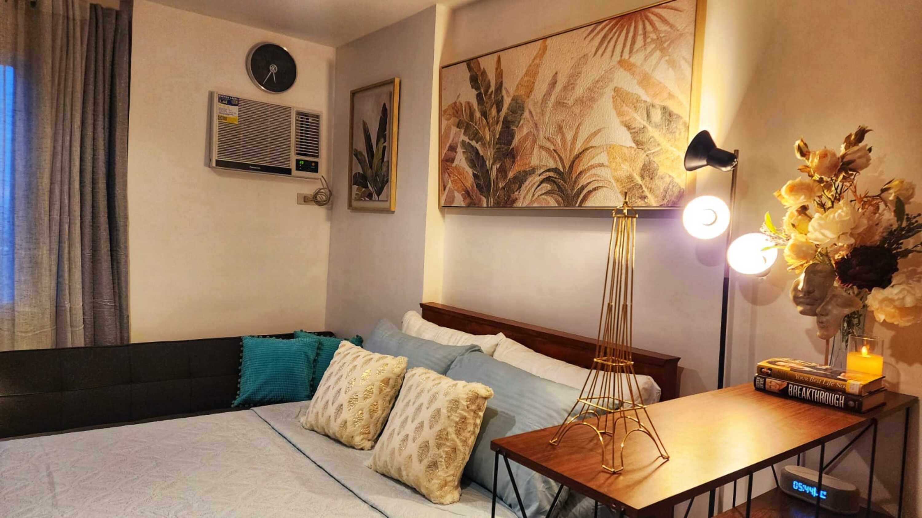 Camera d'albergo con un letto, un comodino con lampada, una sveglia, un quadro e una finestra con tende.
